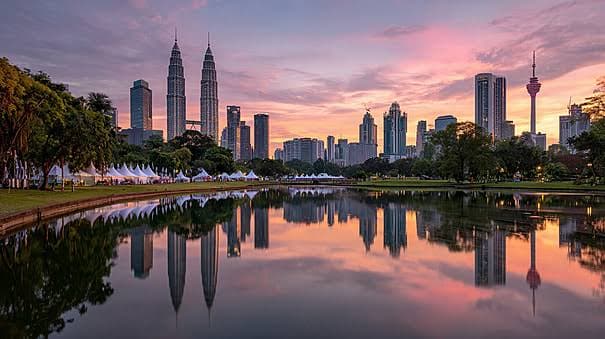 The Kuala Lumpur skyline at sunset, with the Petronas Towers and the KL Tower reflected in the still water of a lakeside park.